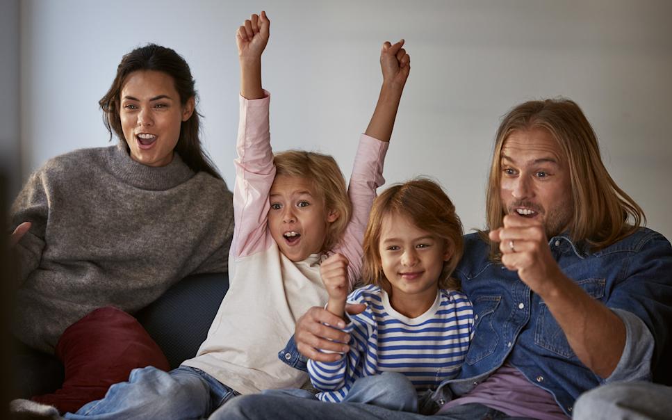 En glad familj som sitter i soffan och tittar på tv, skrattar och hejar.