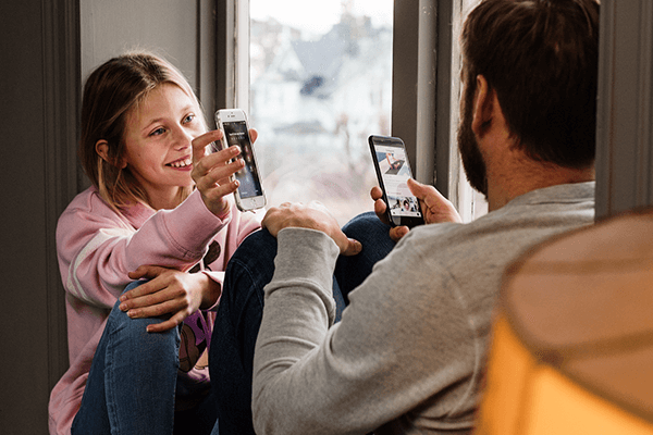 Pappa och barn sitter i ett fönster med varsin mobil.