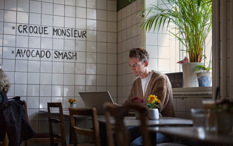 Man på café som använder en dator sittande vid ett bord framför ett fönster med grön växt.