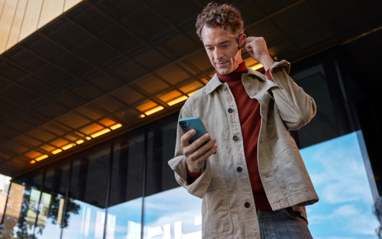 Person med brun skjorta och röd tröja tittar ner i sin telefon samtidigt som han sätter på sig hörlurar.