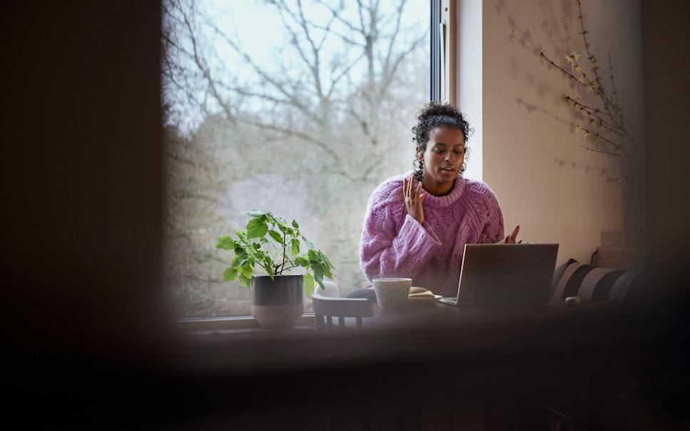 En kvinna sitter vid ett fönster framför sin laptop och gestikulerar med händerna samtidigt som hon pratar.