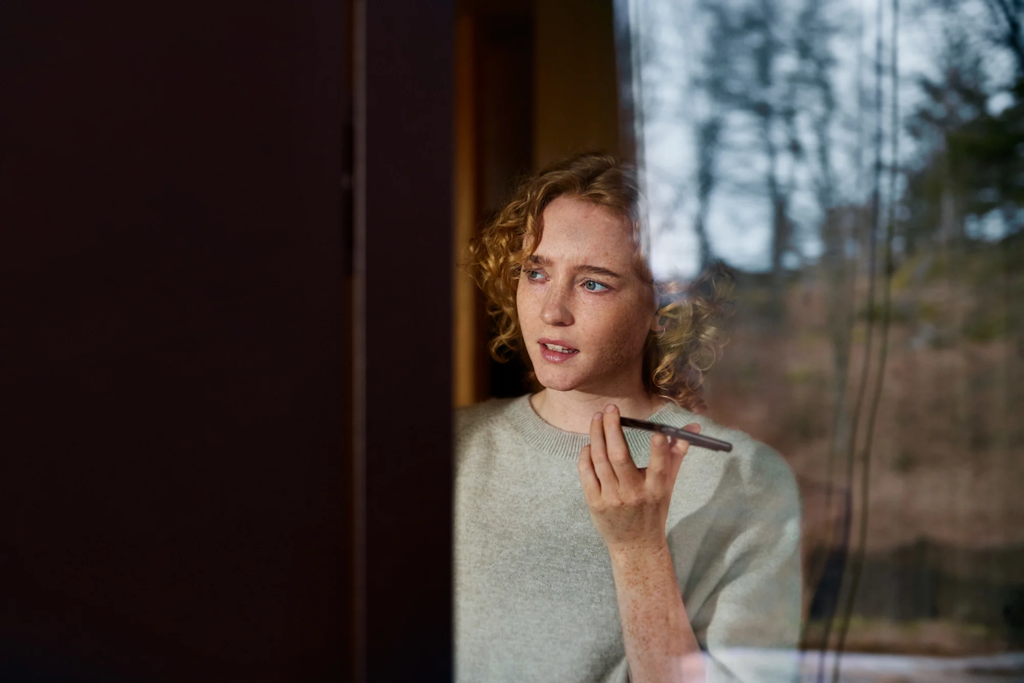 En kvinna håller i en mobil och tittar ut genom fönstret.