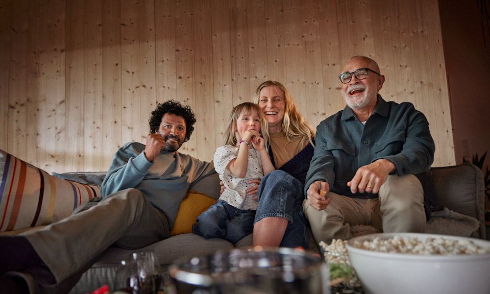 En familj med tre generationer sitter tillsammans i soffan och tittar på tv och skrattar med en stor popcornskål på tv-bordet framför sig.