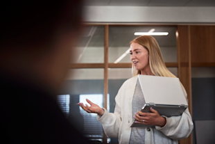 Ung, blond kvinna på kontor pratar med laptop i handen.
