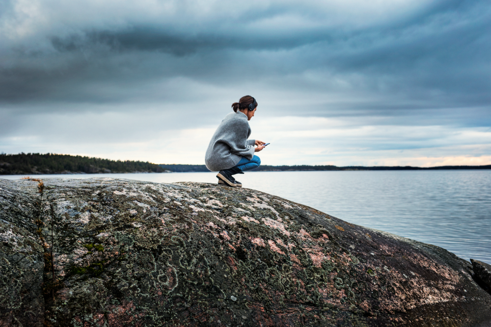 Kvinna med mobil och hörlurar på klippa vid vatten
