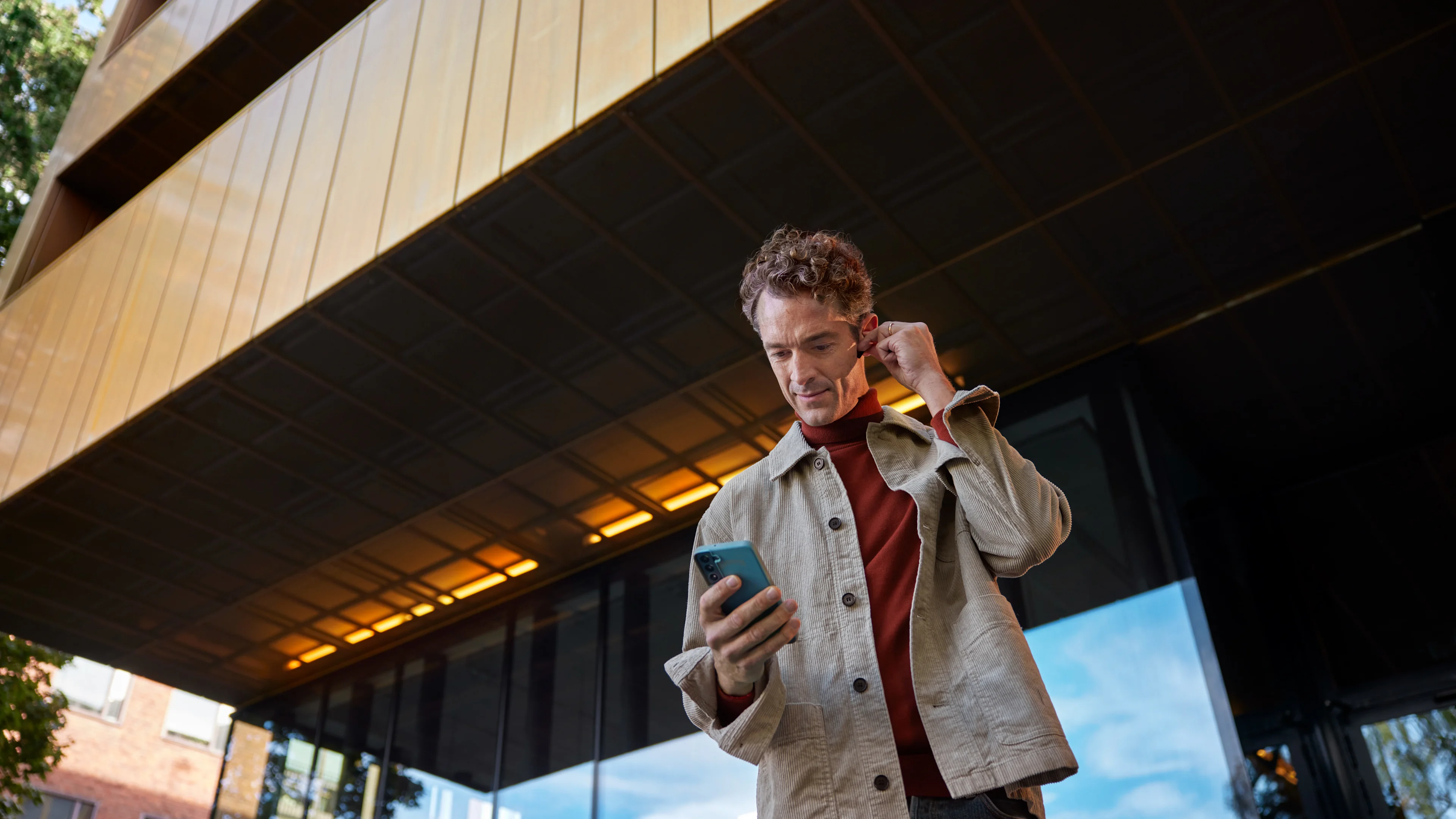 Man framför kontorsbyggnad som tittar på mobil och justerar hörlur.