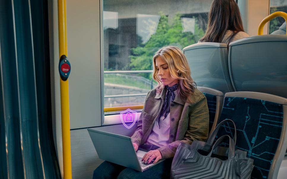 En kvinna sitter i kollektivtrafiken och arbetar på en laptop, ikonen för Trygghetspaketet syns ovanför datorn.