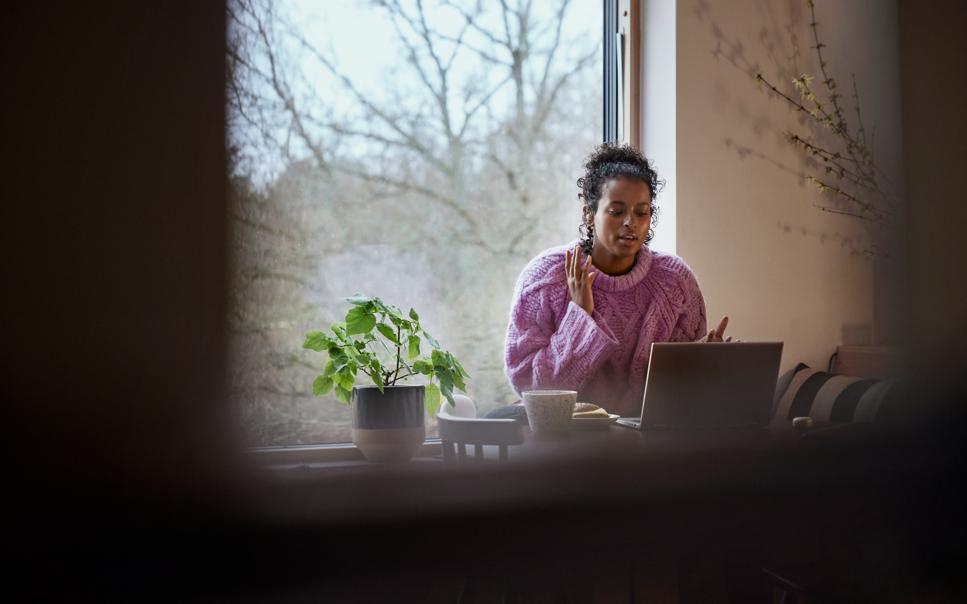 En kvinna sitter vid ett fönster framför sin laptop och gestikulerar med händerna samtidigt som hon pratar.