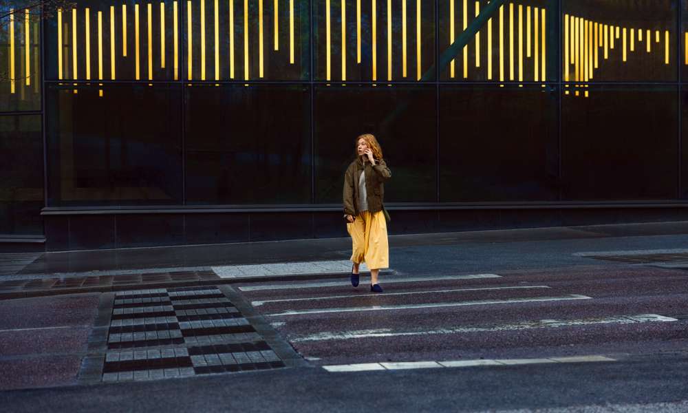 Ung kvinna med gul kjol går över övergångsställe i stad och pratar i mobiltelefon