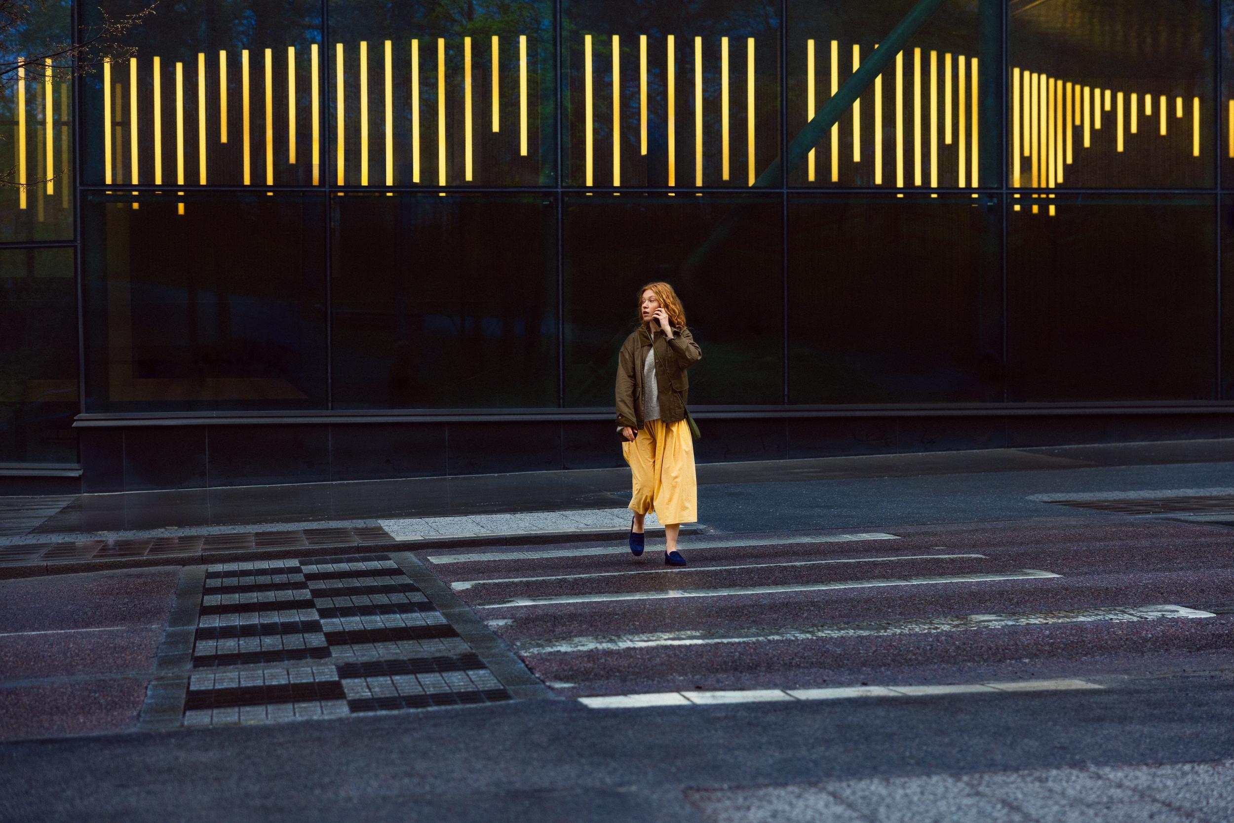 Ung kvinna med gul kjol går över övergångsställe i stad och pratar i mobiltelefon