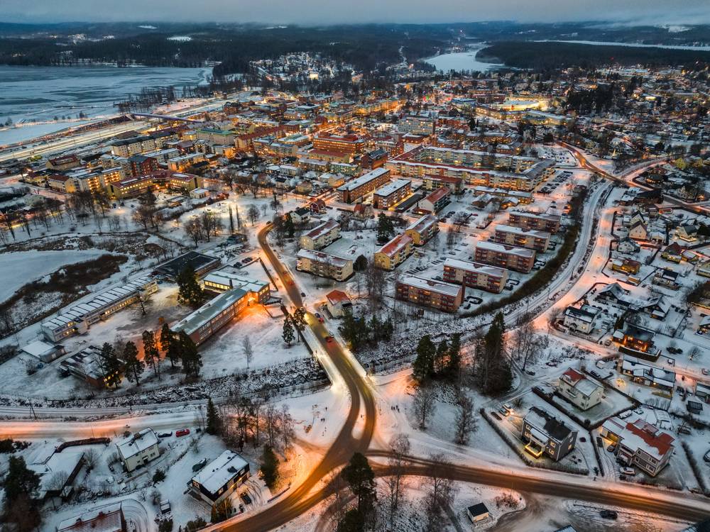 Översiktsbild av ett samhälle under de mörka timmarna av vinterhalvåret. Byggnaderna är upplysta och på gatorna mellan husen syns vägar täckta av snö.