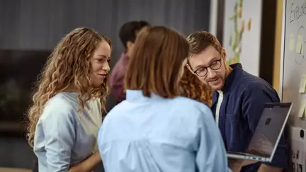 Två kvinnor och en man står upp tittar på en laptop