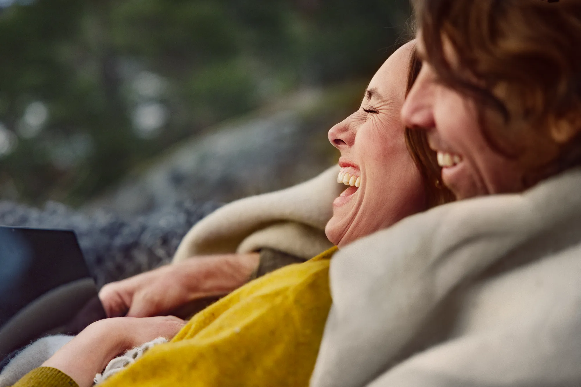 En kvinna och en man sitter omvirad i en filt och tittar på surfplatta tillsammans och skrattar.