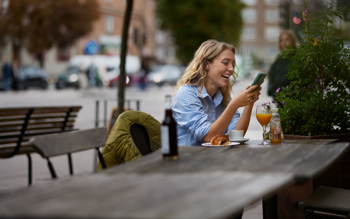 Kvinna utomhus vid ett kafébord tittar på mobilen och skrattar