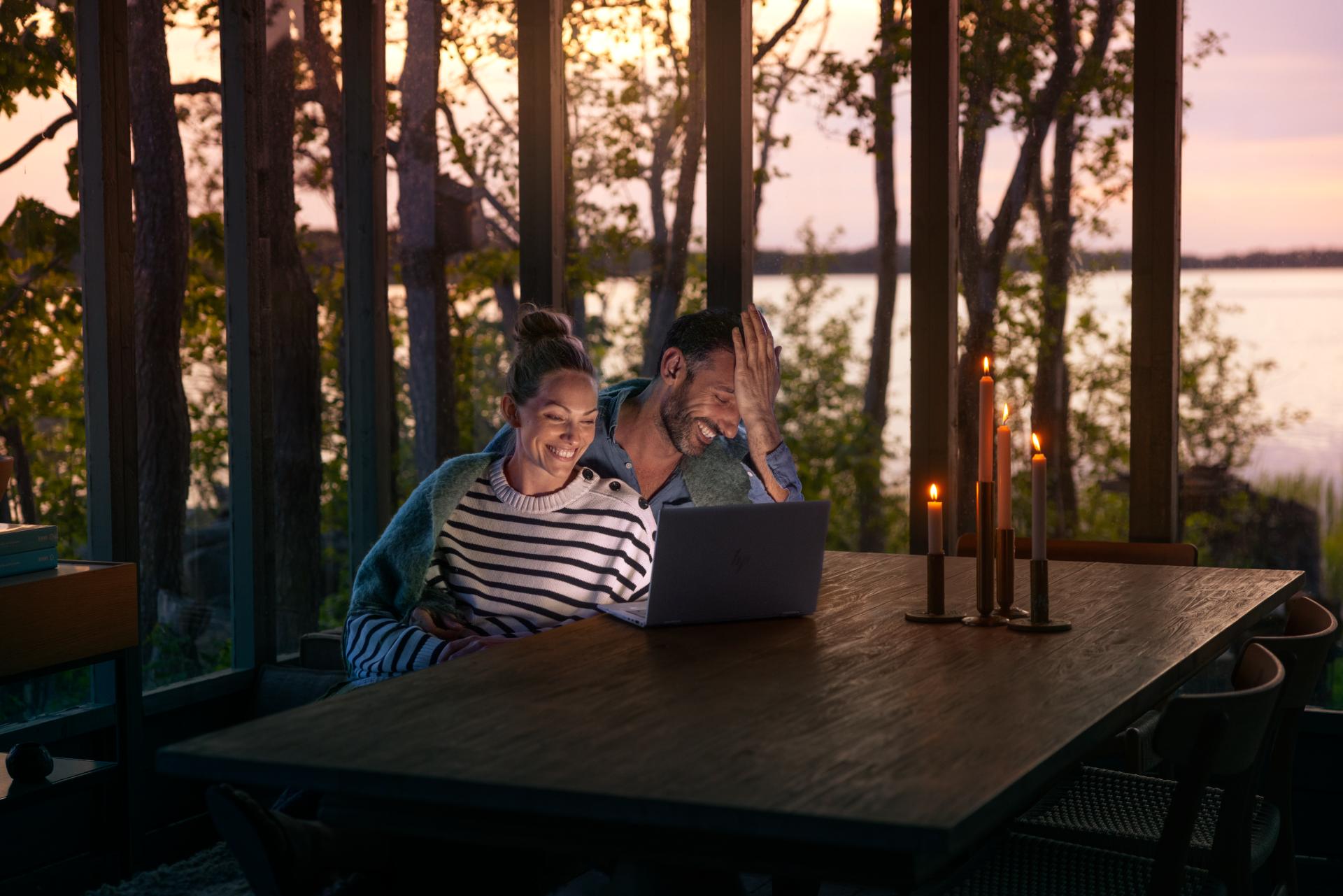 En man och en kvinna sitter vid bordet med levande ljus och tittar på en laptop tillsammans och skrattar med en idyllisk skärgårdsmiljö bakom sig.