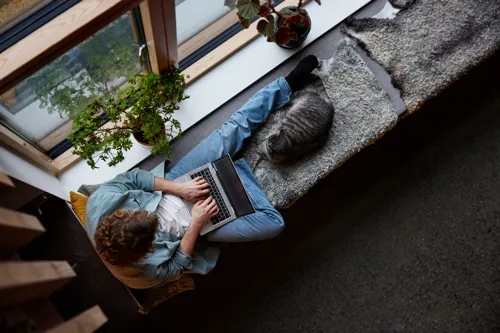 En man med en laptop i knät halvsitter vid ett fönster, bredvid honom ligger en katt.