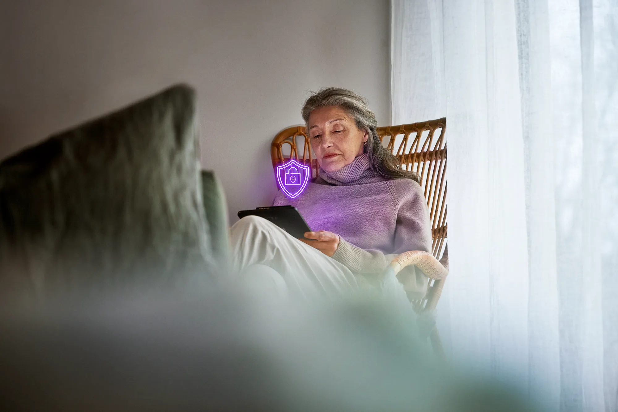 Äldre dam som sitter i en fåtölj och lutar sig tillbaka med sin surfplatta i handen, med en lila säkerhetssköld som representerar Telia Säkert framför.