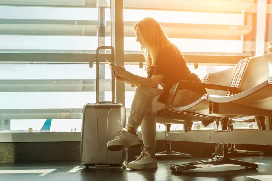 Kvinna med resväska sitter på en flygplats och kollar sin mobil.