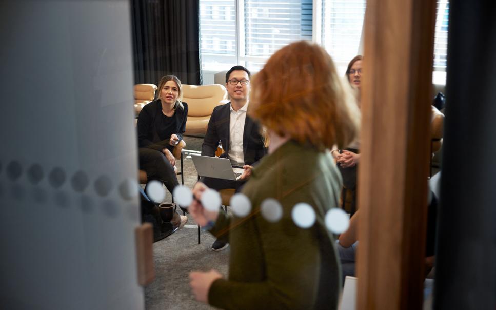 Kvinna i konferensrum sedd genom glasdörr presenterar för kollegor.