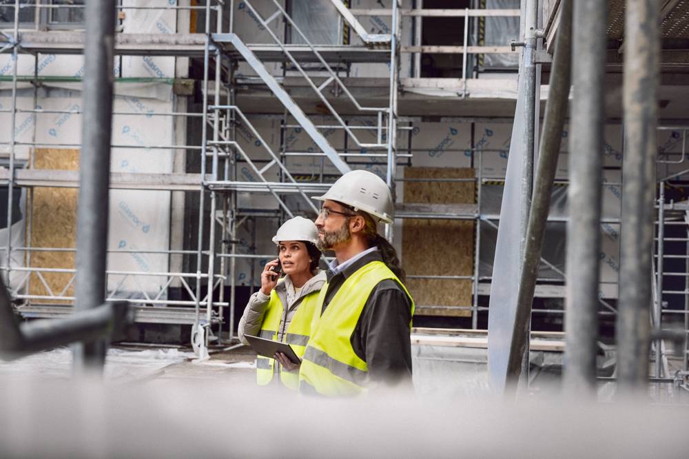 Man och kvinna i bygghjälm och varselväst med iPad på byggarbetsplats.
