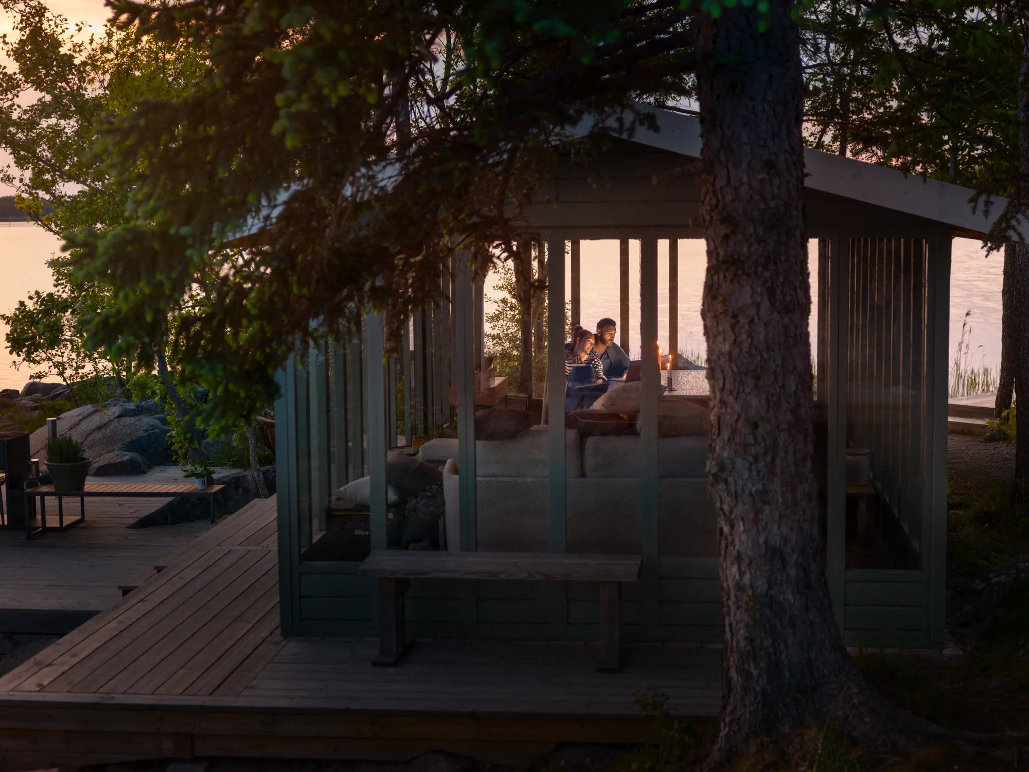 En man och en kvinna som sitter i en mysig stuga precis vid vattnet, som symboliserar Telia Play och Telia tv-paket ger underhållning och bekvämlighet även på avlägsna platser.