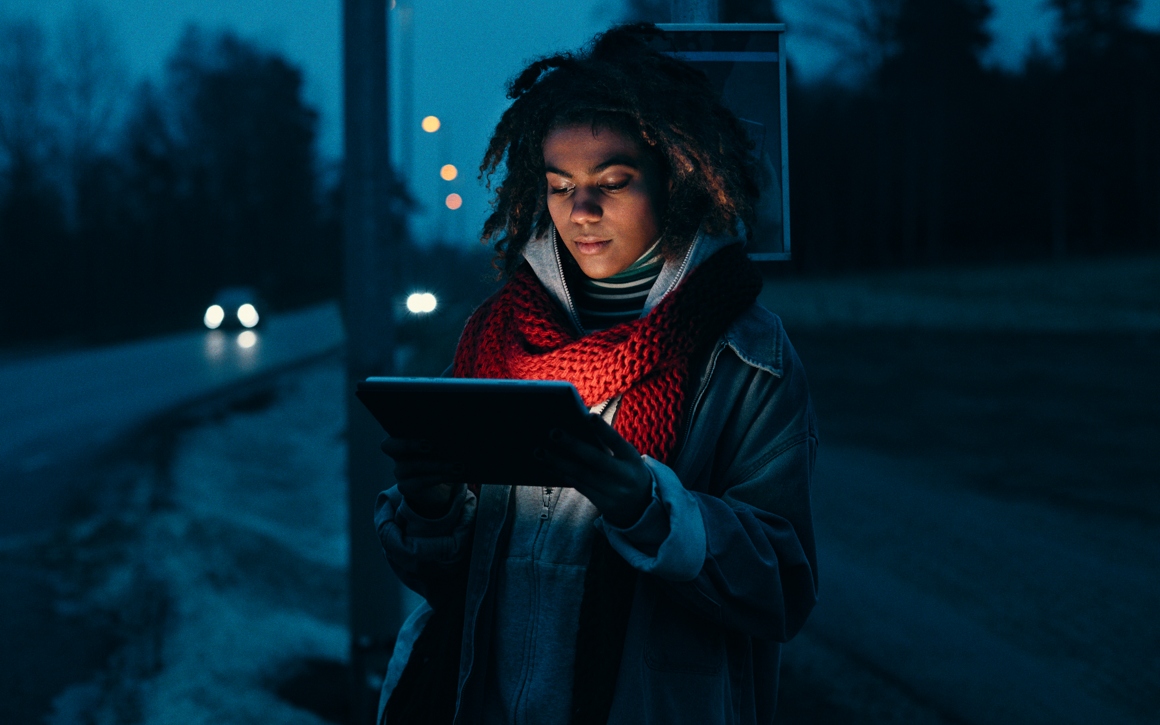 Kvinna står vid busstation på kvällen med surfplatta