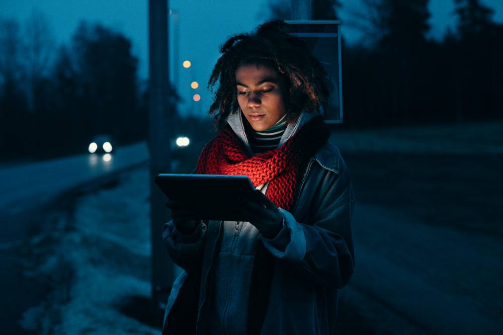 Kvinna står vid busstation på kvällen med surfplatta