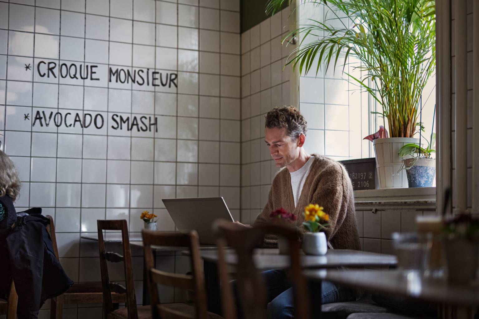 Man på café som använder en dator sittande vid ett bord framför ett fönster med grön växt.