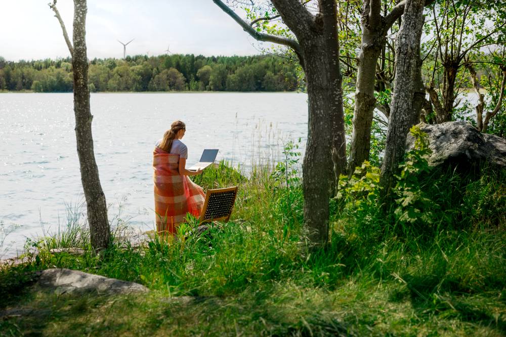 En kvinna står omvirad med en filt runt sig och sin laptop i handen, i en grönskande miljö precis vid vattnet – som symboliserar vårt mobila bredband som låter dig surfa var du vill.