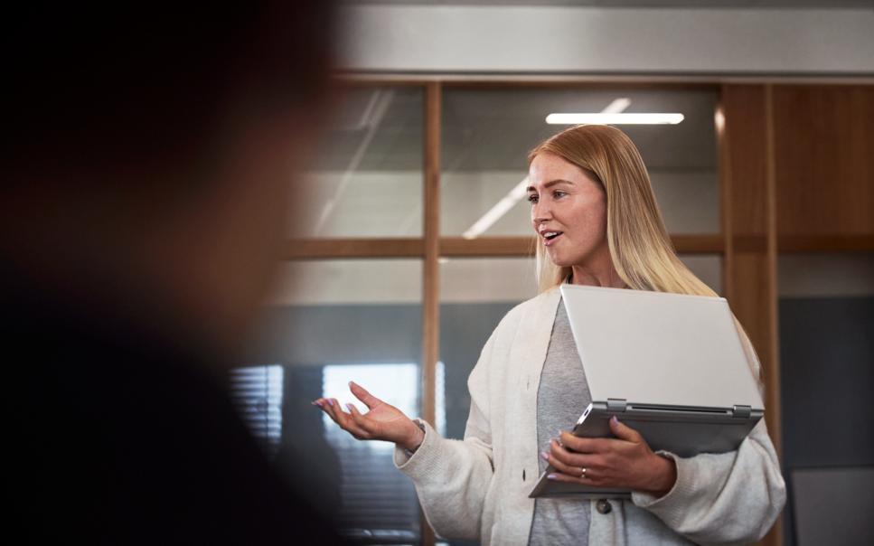 Kvinna står med laptop i famnen och pratar på ett kontor i konferensrum.
