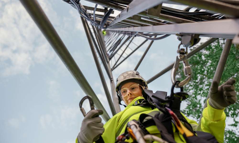 Kvinnlig arbetare i bygghjälm, arbetskläder och skyddsglasögon klättrar i telemast