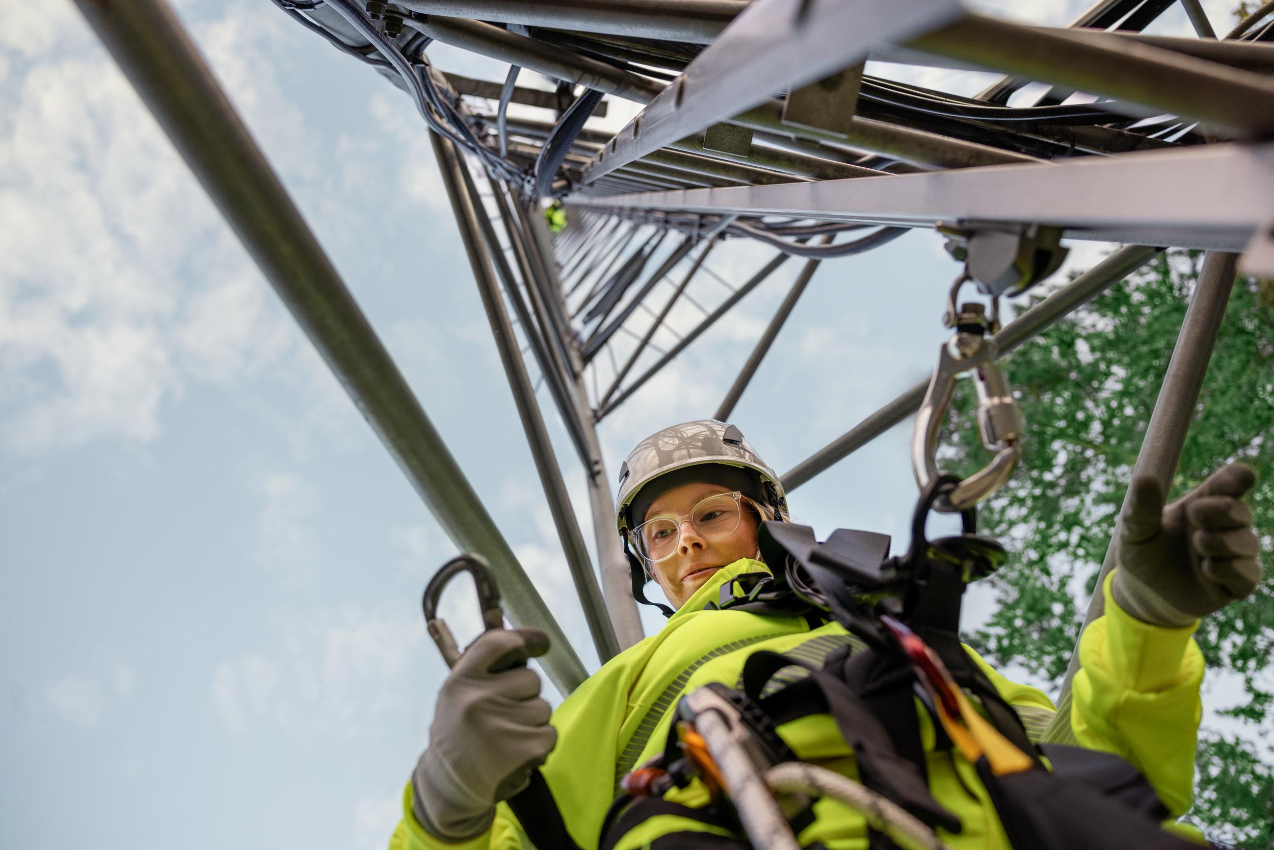 Kvinnlig arbetare i bygghjälm, arbetskläder och skyddsglasögon klättrar i telemast