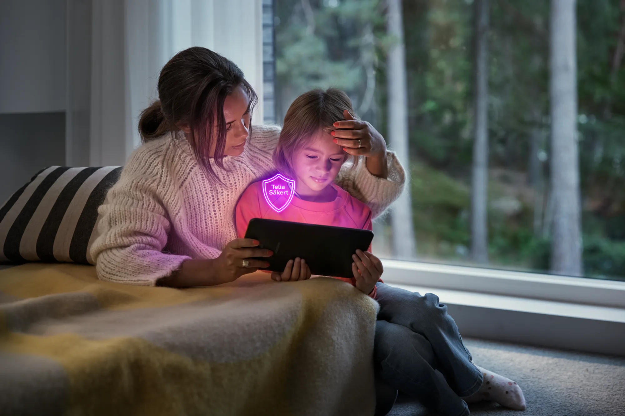 En kvinna och ett barn tittar på en surfplatta, med en lila säkerhetssköld framför med texten "Telia Säkert".