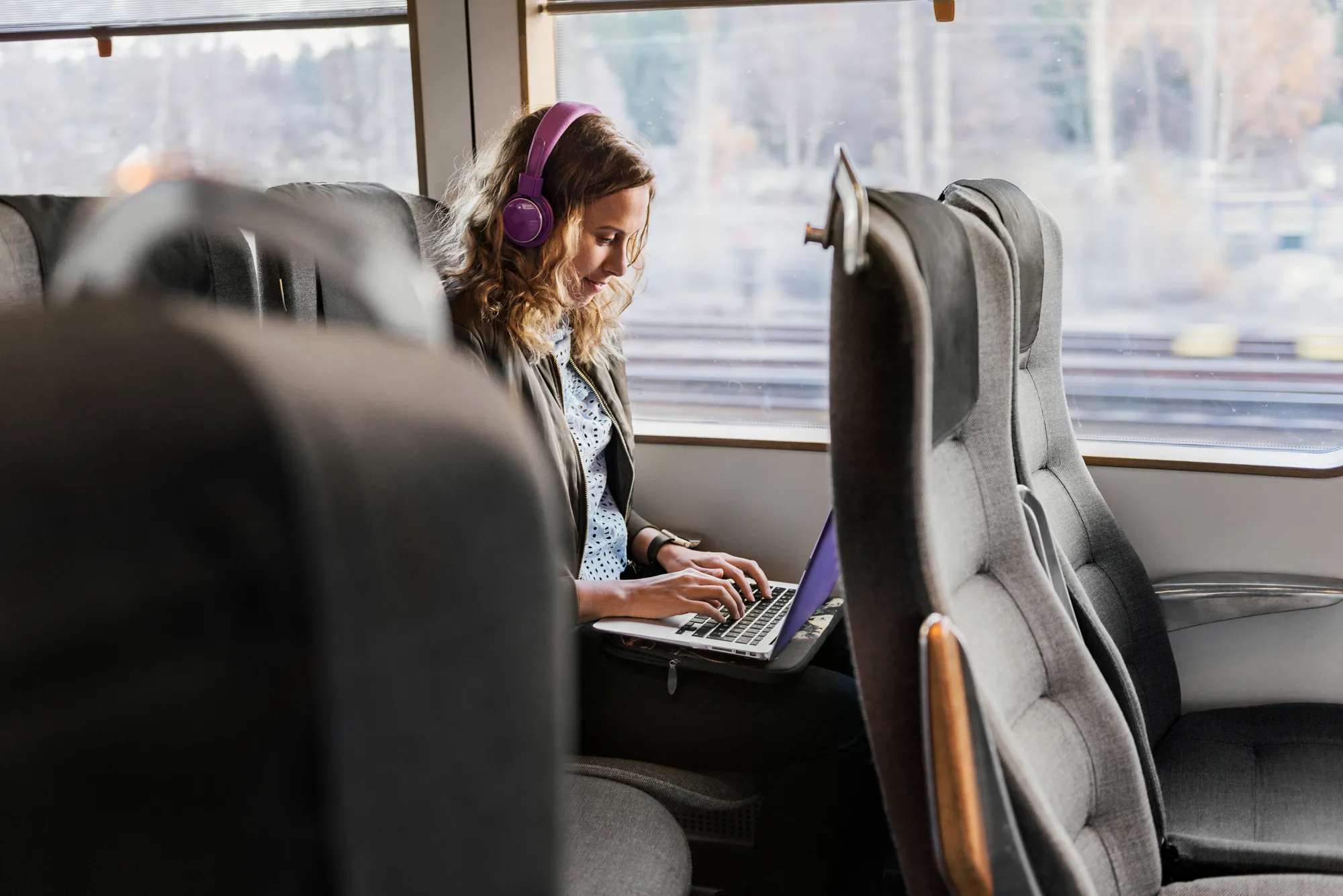 En kvinna med hörlurar sitter på ett tåg och jobbar på en laptop.