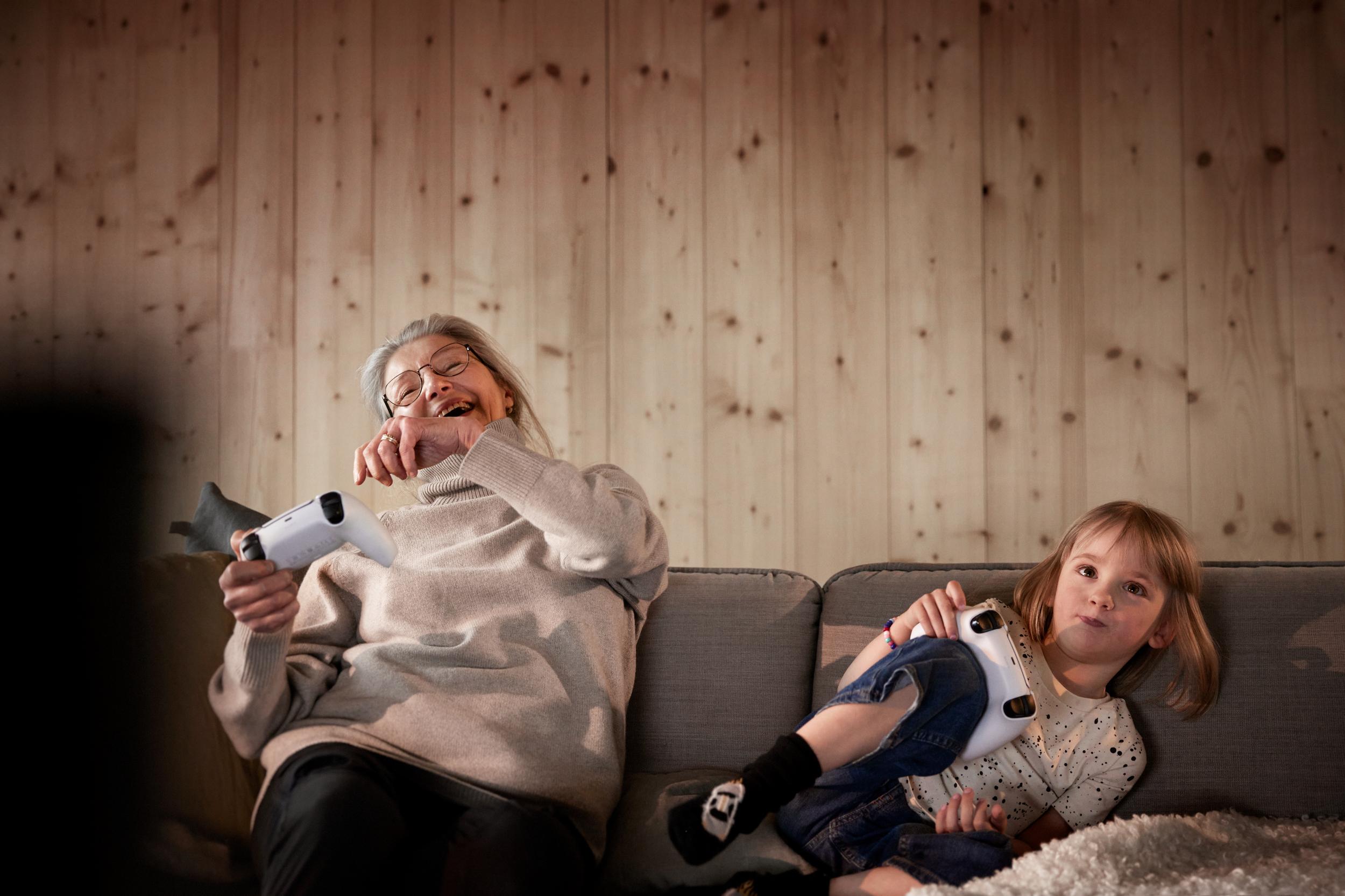 En glad äldre kvinna som sitter och spelar videospel i soffan med sitt barnbarn.