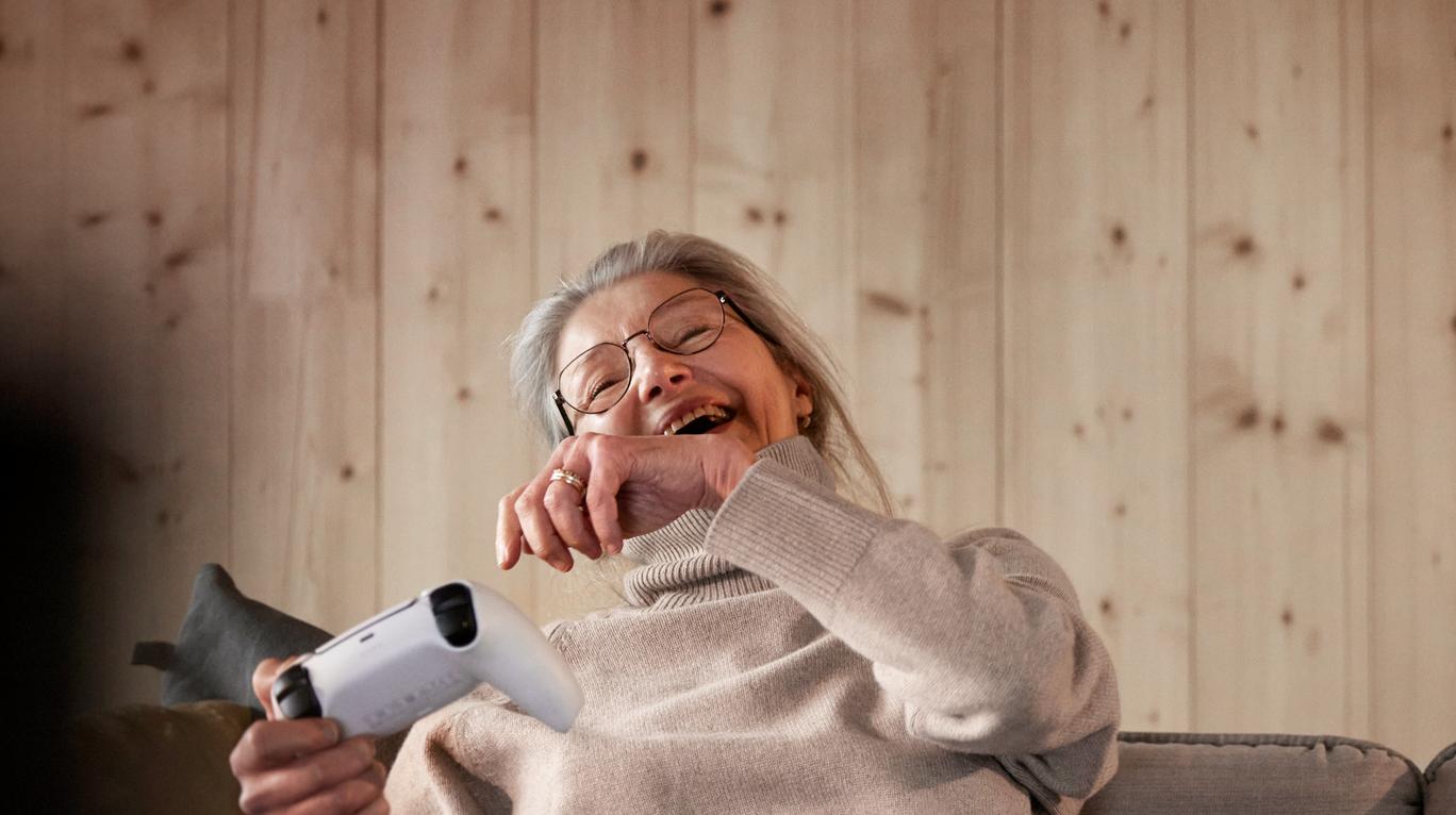 En glad äldre kvinna som sitter och spelar videospel i soffan med sitt barnbarn.