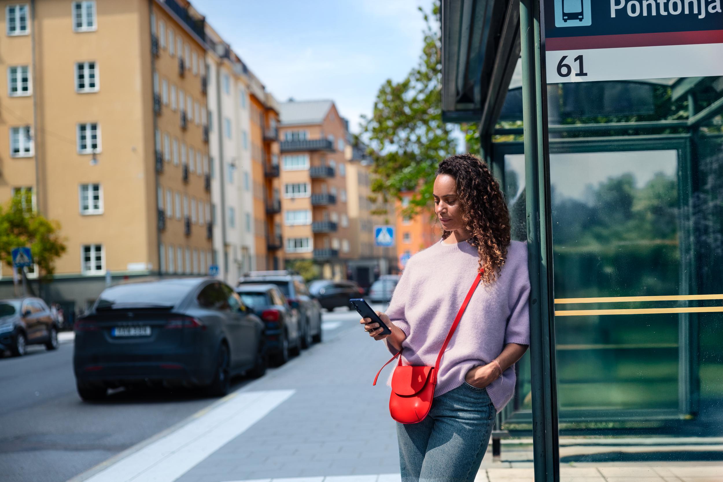 Kvinna vid busshållplats tittar på sin mobil.