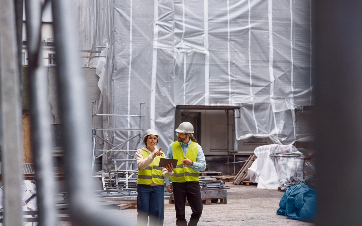 Man och kvinna i varselvästar och hjälmar tittar på platta på byggarbetsplats