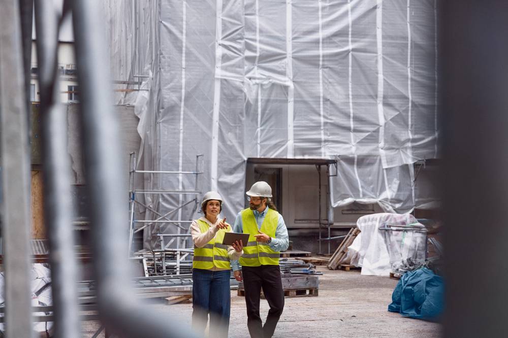 Man och kvinna i varselvästar och hjälmar tittar på platta på byggarbetsplats