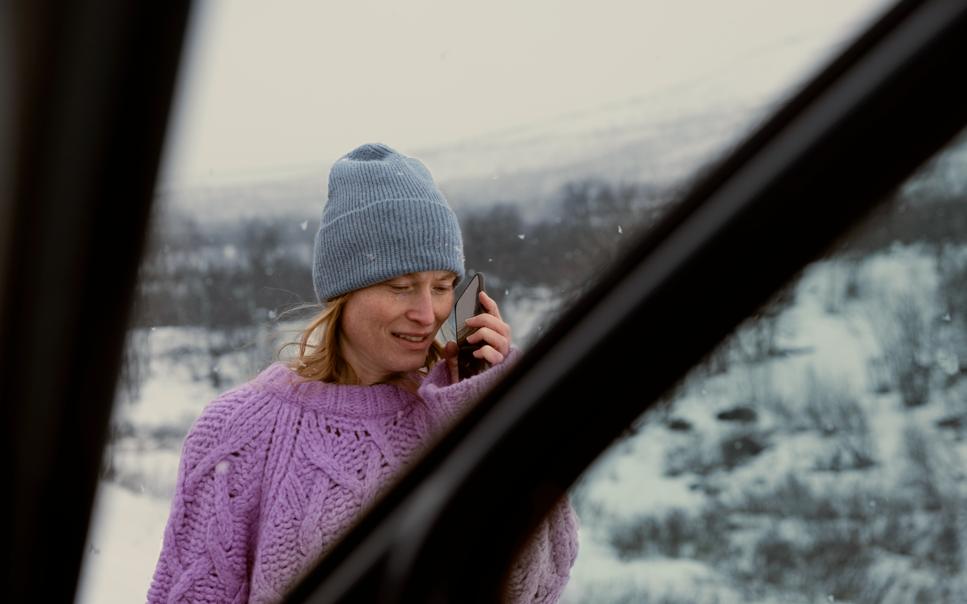 Kvinna med mössa pratar i mobil utanför bil i snöigt landskap.