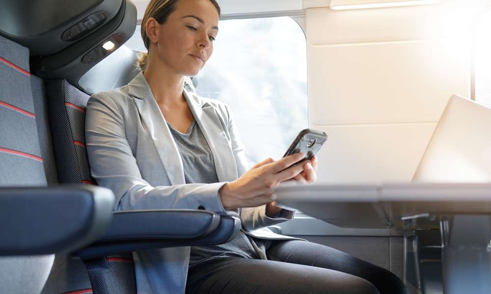 Kvinna på affärsresa på tåg, tittar på mobil och har laptop framför sig.