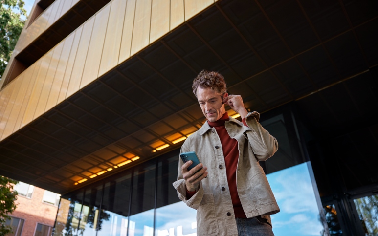 Person med brun skjorta och röd tröja tittar ner i sin telefon samtidigt som han sätter på sig hörlurar.