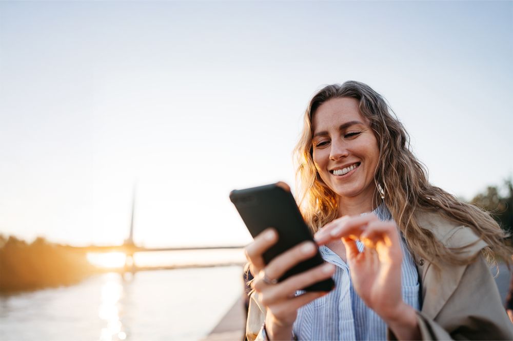 Kvinna tittar leende på mobiltelefon framför bro och vatten, soligt.