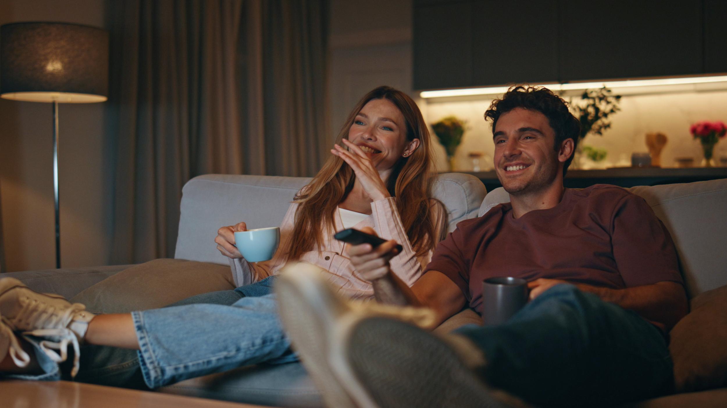 Ungt par sitter i soffan, tjejen med en tekopp i handen och killen en fjärrkontroll för att välja vad de ska streama.