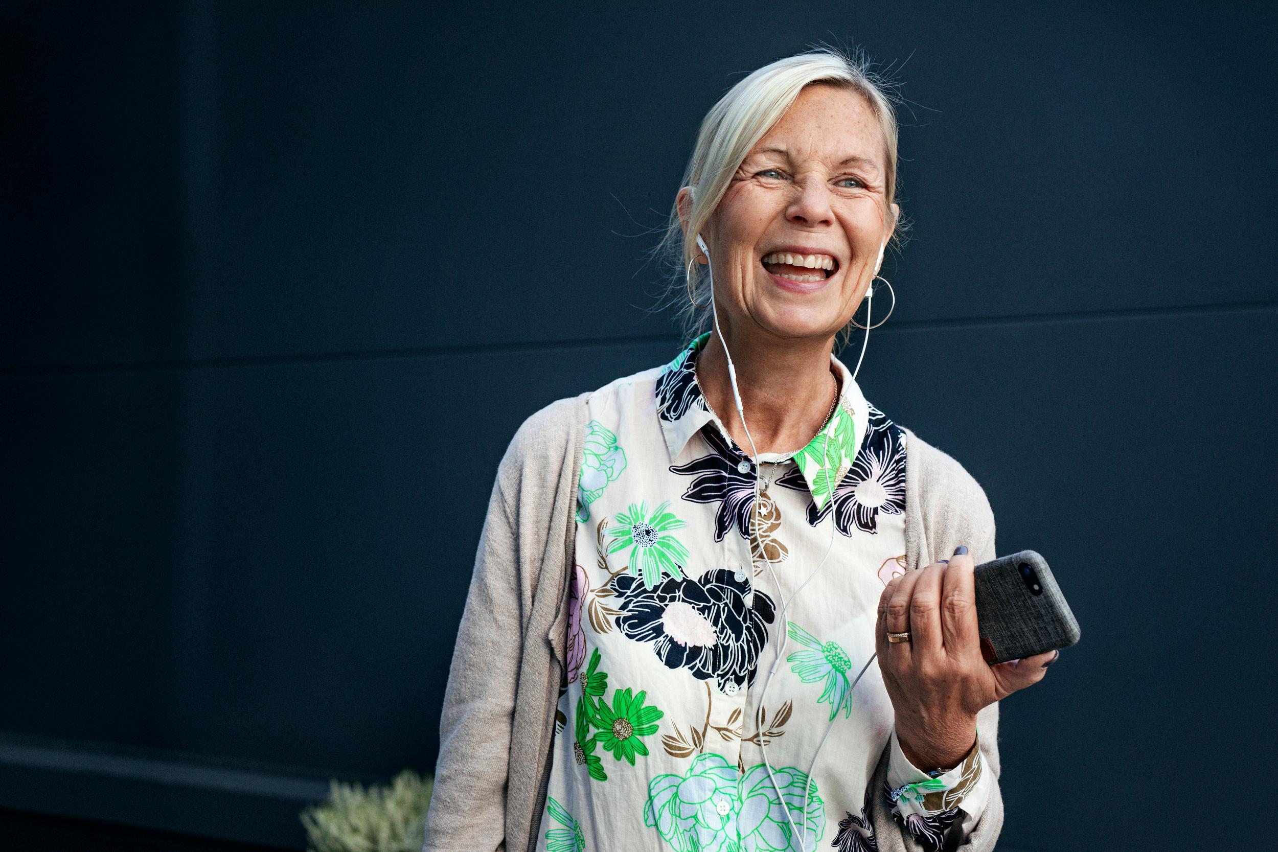 En äldre kvinna som ler och skrattar, med hörlurar i öronen och mobilen i handen.
